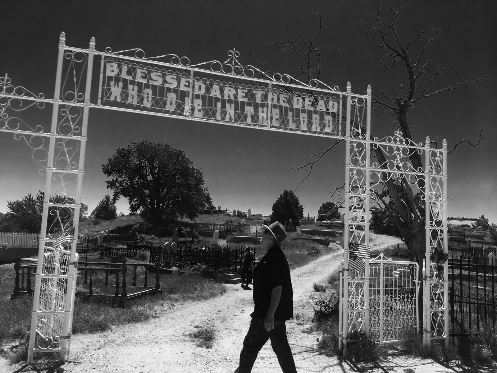 Black-and-white photo of a man in a hat walking past a white iron cemetery gate arched with the words Blessed are the dead who die in the Lord