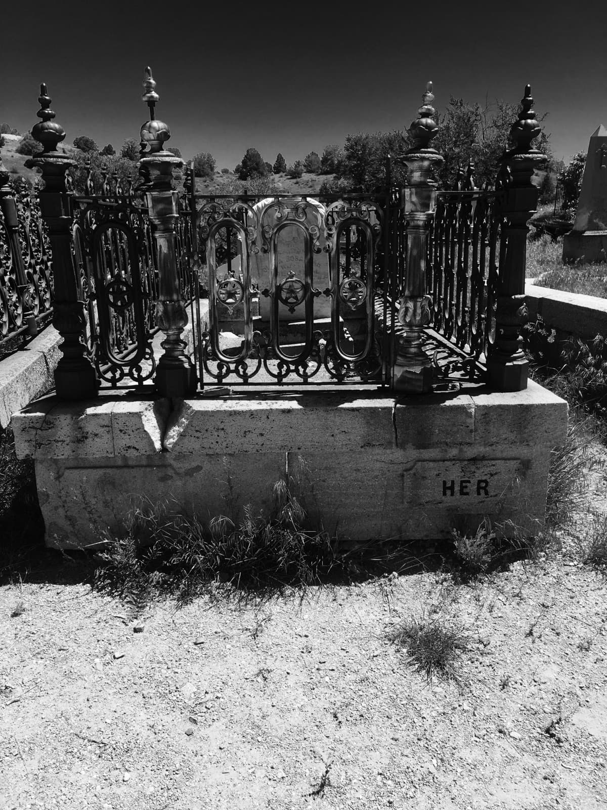 Black-and-white photo of an iron-fenced grave plot on a stone base in a hillside cemetery