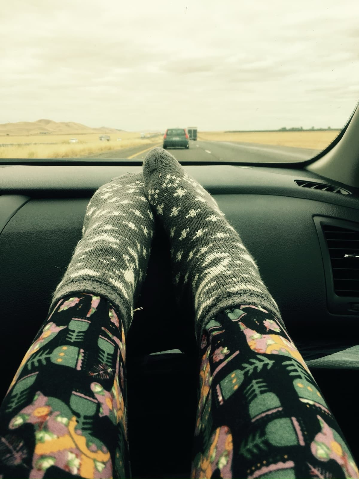 A passenger's feet in patterned leggings propped on a car dashboard, an open road ahead