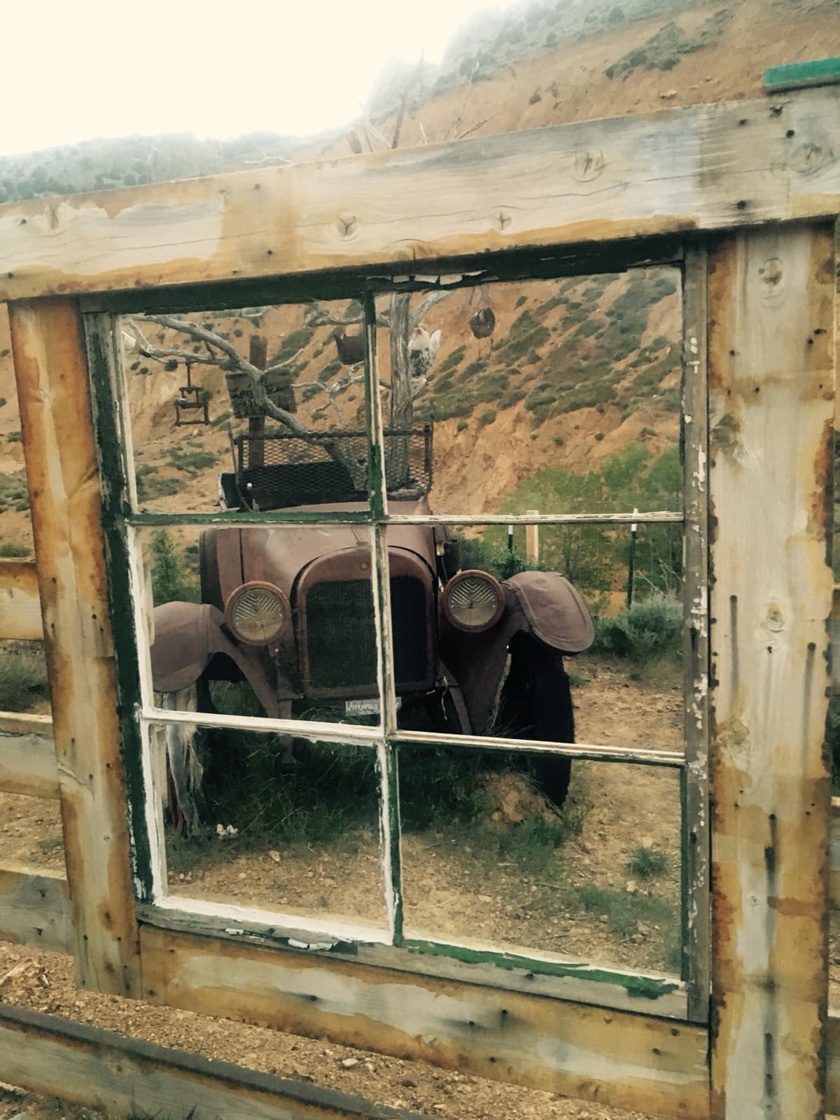 An old rust-brown car seen through a weathered window frame, a dry hillside beyond