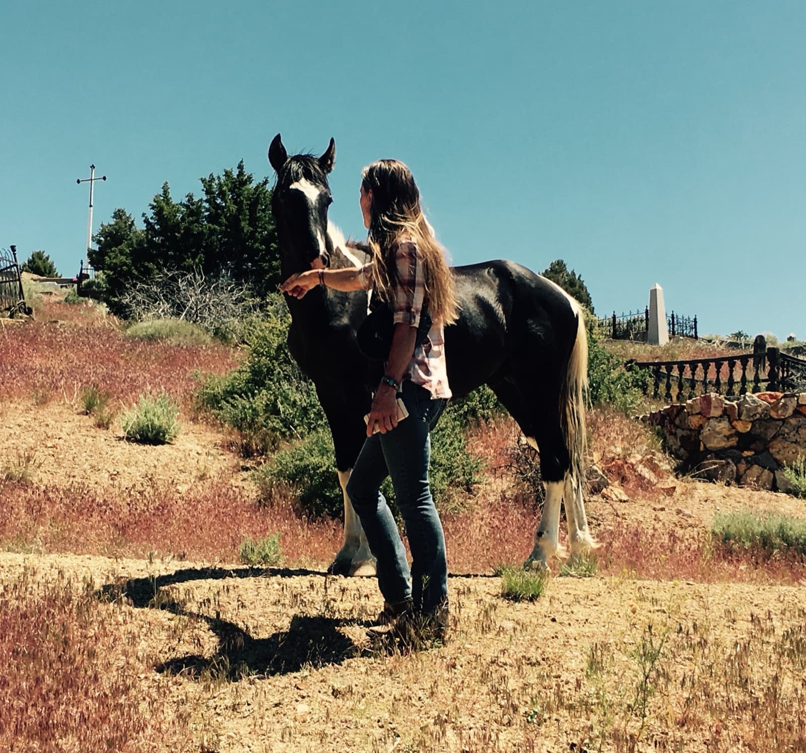 A woman in a plaid shirt stroking the face of a black-and-white paint horse on a dry hillside, a cemetery fence and obelisk behind