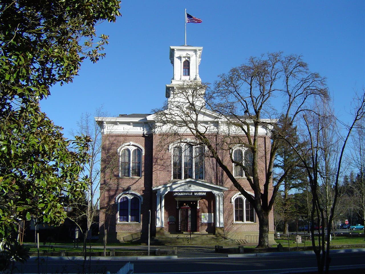 The historic brick Jacksonville Museum with its white cupola and flag, framed by trees under a blue sky, Jacksonville, Oregon