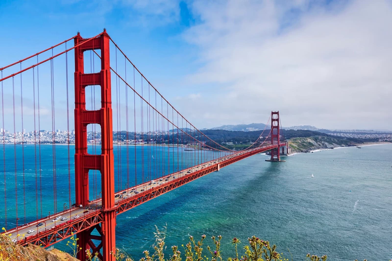 The Golden Gate Bridge's red towers and cables stretching across the bay under a blue sky with scattered cloud, San Francisco skyline behind